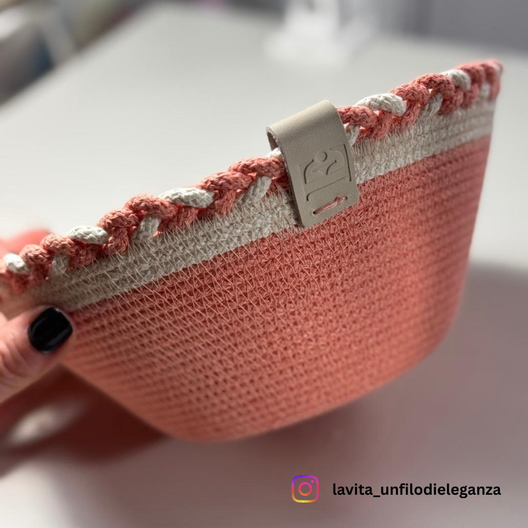 A close-up of a woven basket or tray in coral pink and cream, held by a hand with black nail polish. The basket has a decorative braided trim and a beige leather strap with an embossed logo.
