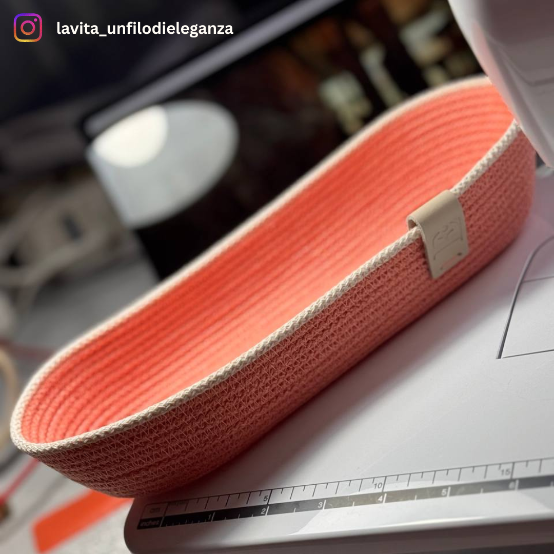 A close-up of a woven oval storage basket in a coral pink and cream color combination, resting on a white sewing machine. The basket has a small beige leather tag on the side.